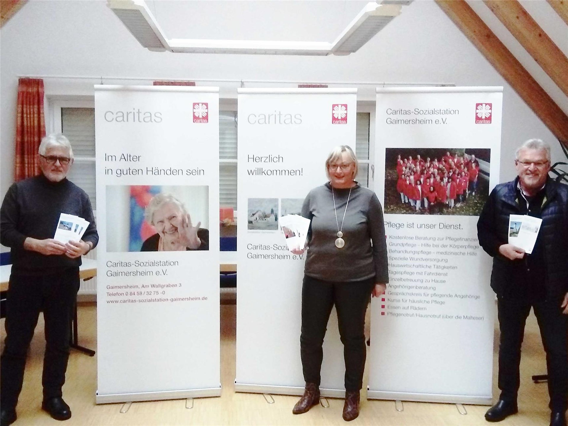Zwei Männer und eine Frau zeigen Prospekte von Caritas-Roll-ups